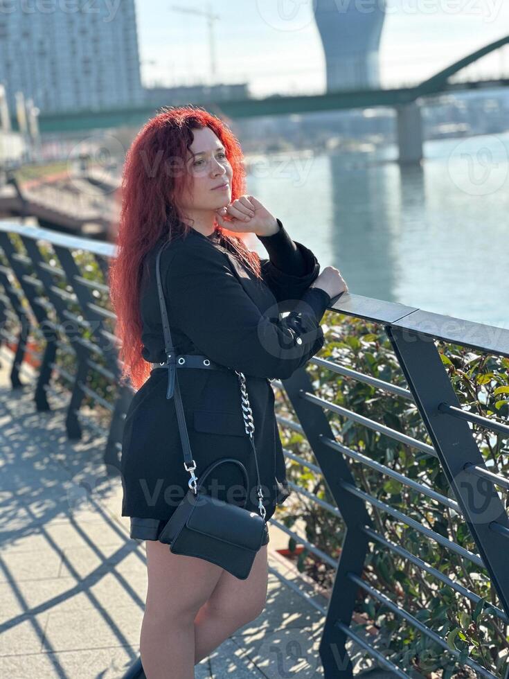 Millennial woman with long curly red hair leans on a black railing, gazing thoughtfully at the camera on a sunny day. Background features modern buildings and a river, evoking a sense of urban calm photo