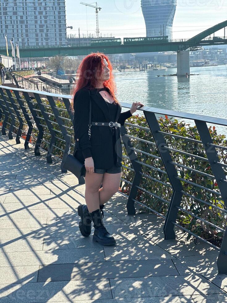 Millennial woman with long curly red hair leans on a black railing, gazing thoughtfully at the camera on a sunny day. Background features modern buildings and a river, evoking a sense of urban calm photo