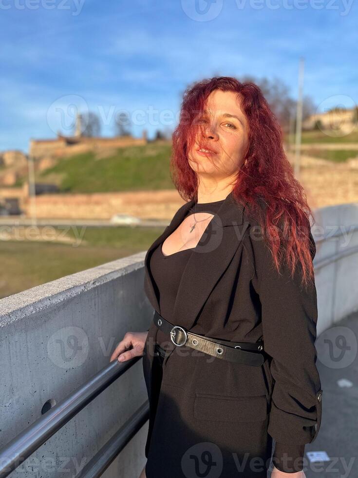 Millennial woman stands confidently in sunlight, with vibrant red hair flowing and dressed in a chic black outfit. Background of a clear blue sky and distant scenery enhances the dynamic photo