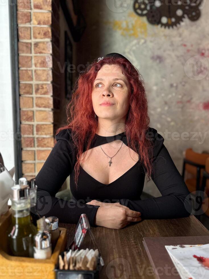 Millennial woman with red hair sits thoughtfully at a rustic cafe table, surrounded by a cozy and artistic atmosphere. Soft lighting and her pensive expression convey photo