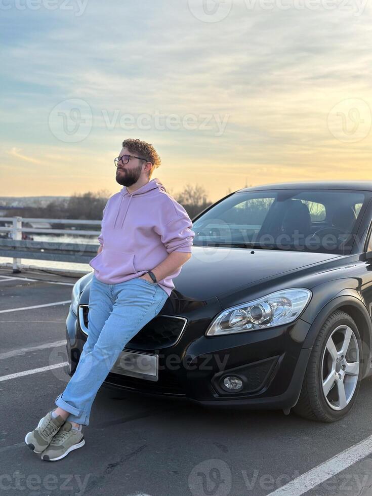 Millennial man in a casual hoodie leans confidently against a sleek black car in a parking lot. Peaceful evening sky forms a serene backdrop to this moment of relaxation and contemplation photo