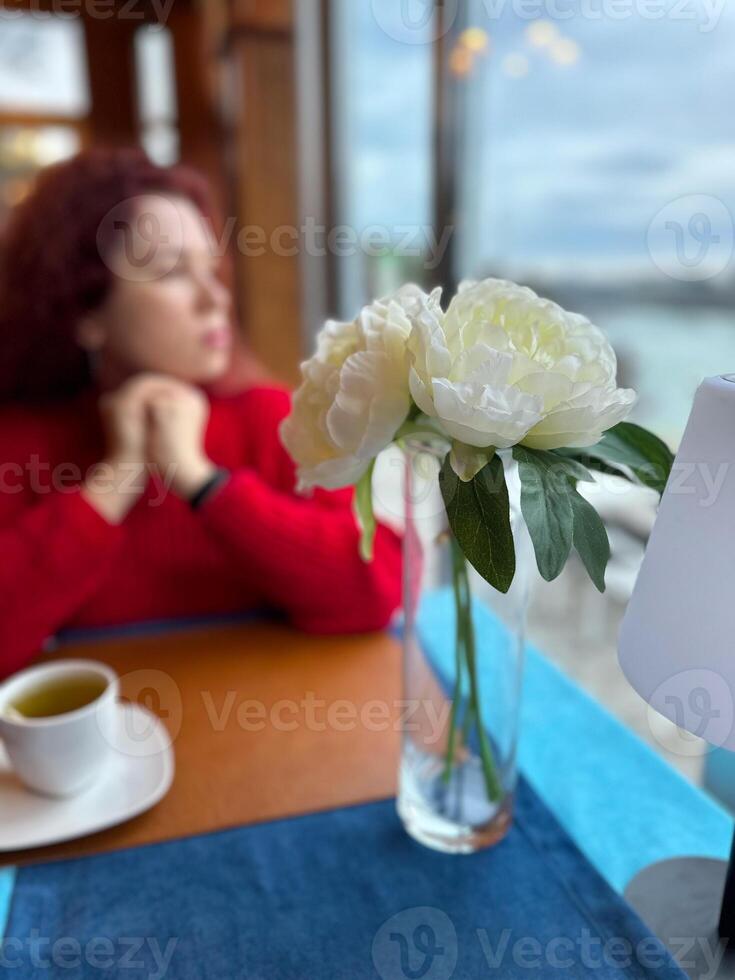 Focus on a delicate white flower in a vase with a blurred woman in background, dressed in red, depicting a serene indoor scene. Contrasting colors create a sense of tranquility and contemplation. photo