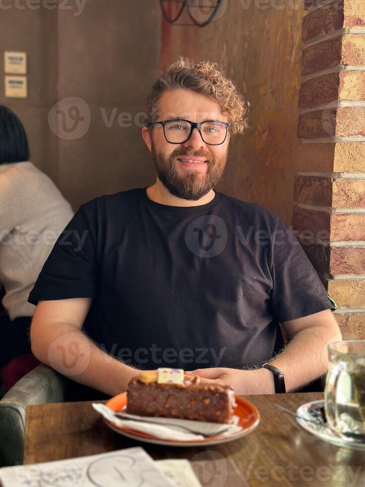 Bearded curly millennial man sitting in a cozy cafe with a slice of cake in front, enjoying a quiet moment in a rustic setting. photo