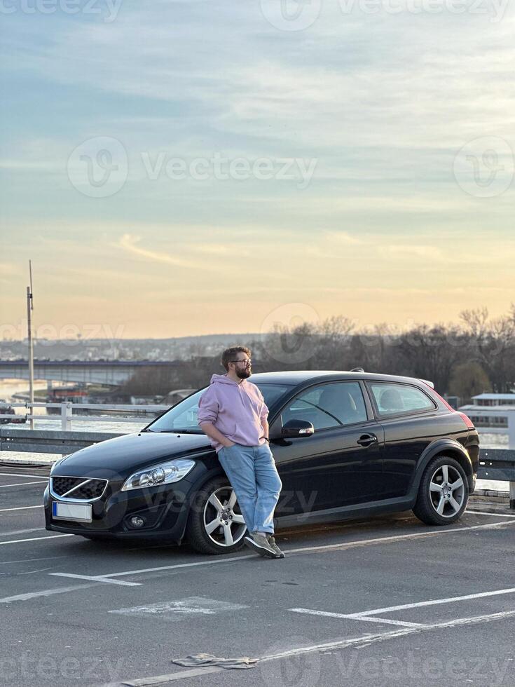 Millennial man in a casual hoodie leans confidently against a sleek black car in a parking lot. Peaceful evening sky forms a serene backdrop to this moment of relaxation and contemplation photo
