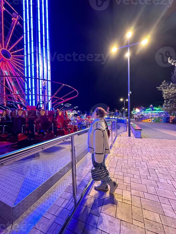 Millennial woman in a fluffy jacket stands illuminated by the colorful lights of a carnival ride in amusement park, gazing upwards with a joyful expression. Winter festive atmosphere photo