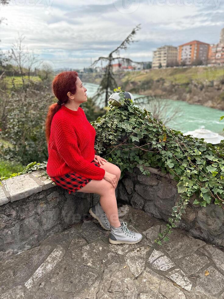 Millennial woman in a red sweater sitting on a stone ledge, enjoying a tranquil river view with a backdrop of city buildings. Autumn scene photo
