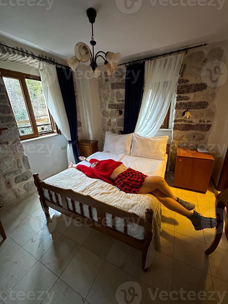 Millennial woman in a red sweater resting on a bed in a rustic bedroom with stone walls and white curtains. Moment of relaxation and comfort captured in a cozy setting in autumn photo