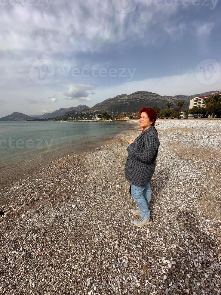 Millennial woman stands alone on a pebbled beach, gazing out at the tranquil sea under a partly cloudy sky. the serene setting is framed by distant mountains and a coastal cityscape photo