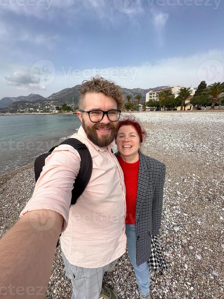 couple takes a cheerful selfie on a pebbled beach, with vast mountains and a serene sea in the background. their relaxed expressions and casual attire complement the picturesque coastal scenery photo