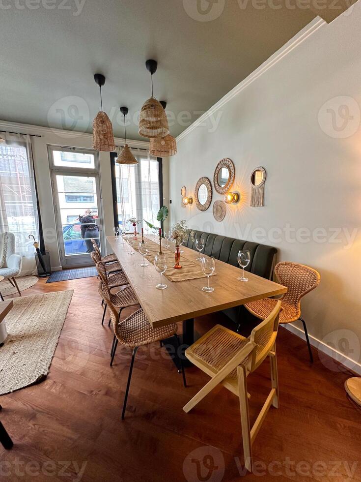 Cozy dining room with a long wooden table is elegantly set with wine glasses and natural decor. wicker chairs and pendant lights create a warm and inviting atmosphere, perfect for a gathering photo