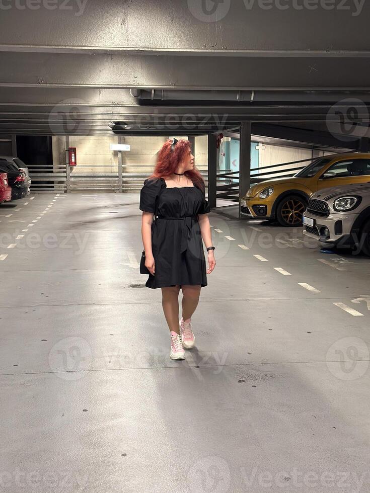 Millennial woman in a black dress walks through an empty parking garage, her pink sneakers adding a pop of color. Setting is urban and modern, with cars lined up neatly in the dimly lit space photo