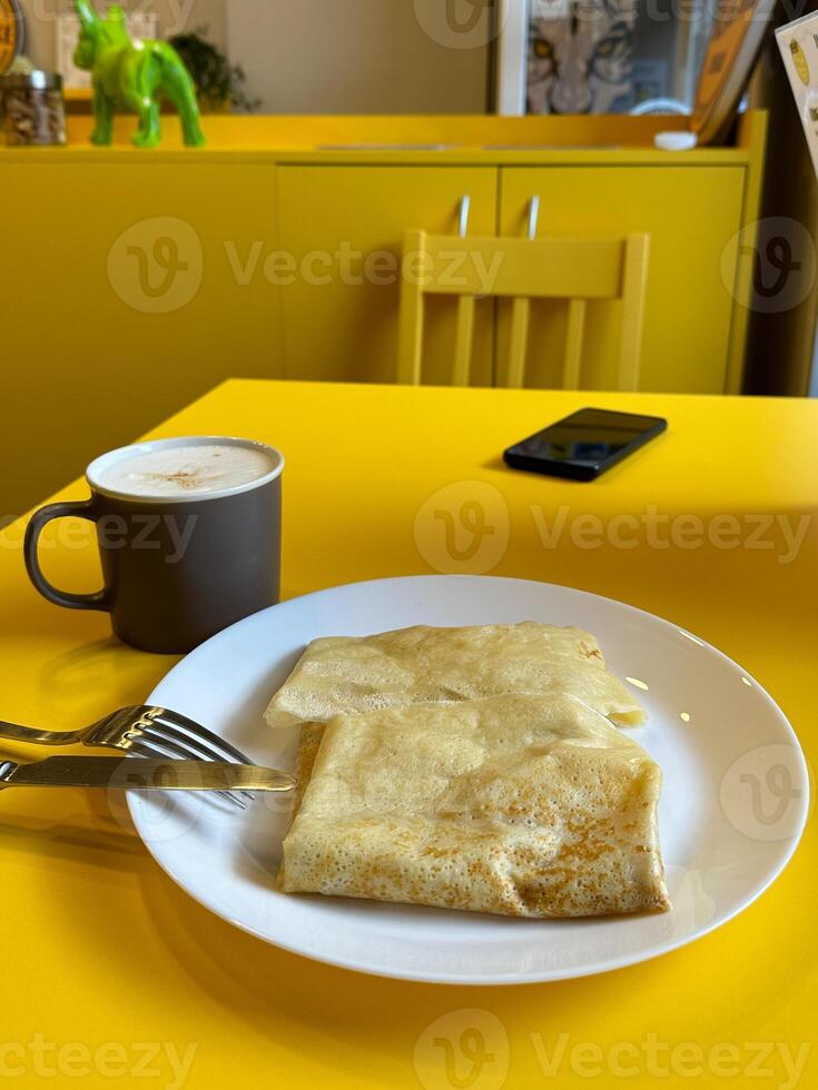 Breakfast setting with a plate of crepes and a frothy cappuccino on a bright yellow table. Cafe with modern design. Pancakes with different fillings. Food concept photo