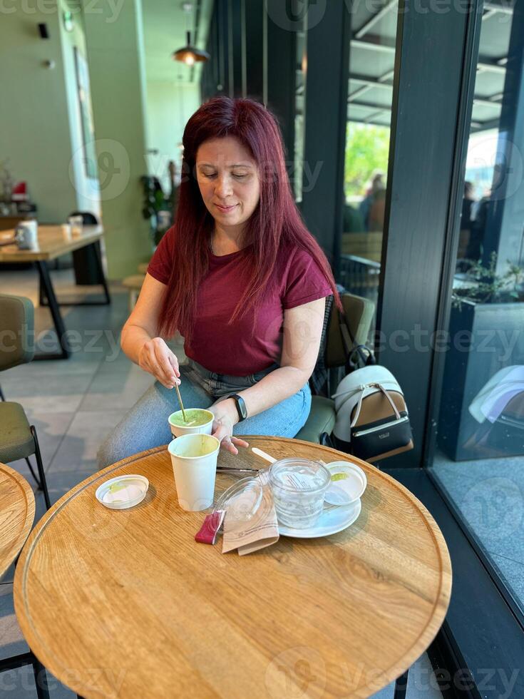 Woman with long red hair enjoys a casual moment at a coffee shop. Girl drink coffee in cafe. Bright interior and large windows create a cozy and inviting atmosphere. Breakfast time photo