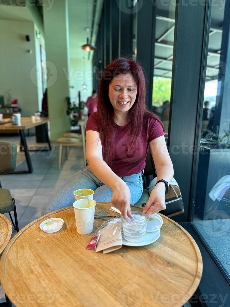 Woman with long red hair enjoys a casual moment at a coffee shop. Girl drink coffee in cafe. Bright interior and large windows create a cozy and inviting atmosphere. Breakfast time photo