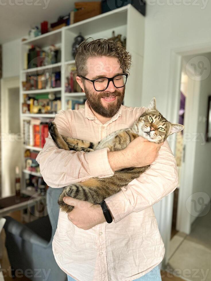 Warm moment between a millennial man and his cat, bond and affection shared in a cozy home environment. The background features a bookshelf at home. Pet and friendship concept photo