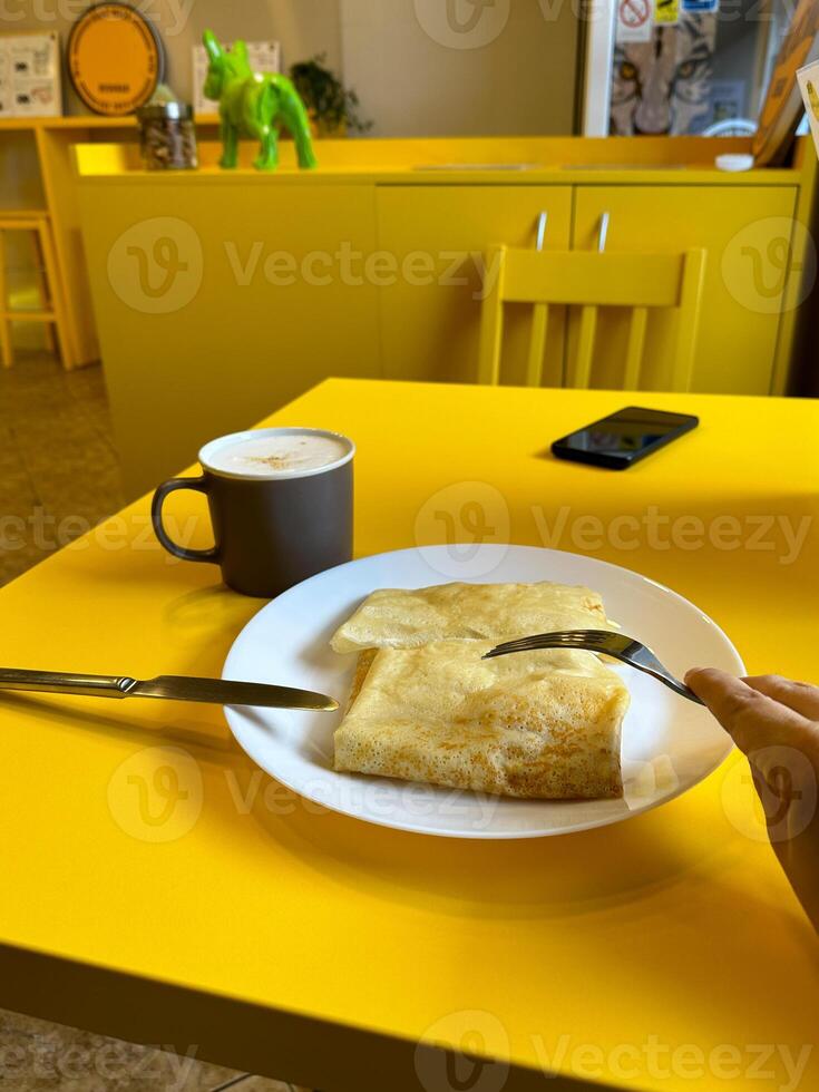 Breakfast setting with a plate of crepes and a frothy cappuccino on a bright yellow table. Cafe with modern design. Pancakes with different fillings. Food concept photo