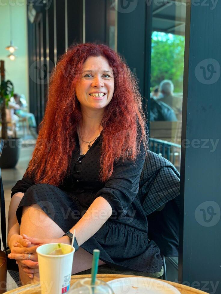 Millennial woman with red hair sits thoughtfully by a large window, gazing outside. Cozy cafe atmosphere, warm and serene mood. photo