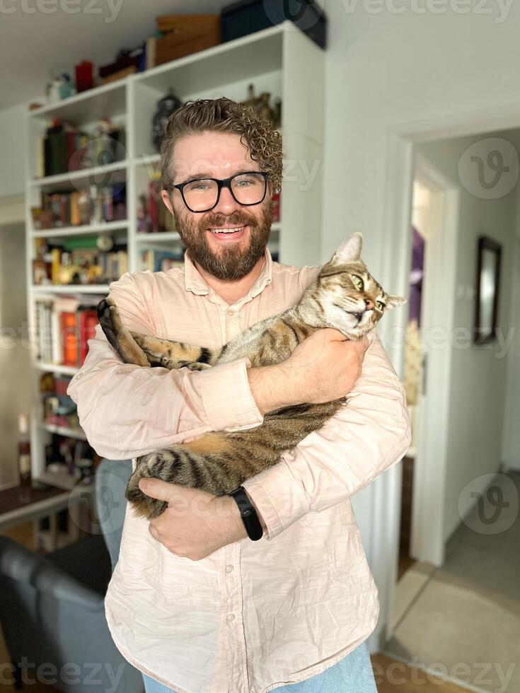 Warm moment between a millennial man and his cat, bond and affection shared in a cozy home environment. The background features a bookshelf at home. Pet and friendship concept photo