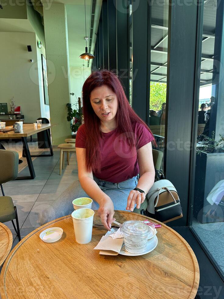 Woman with long red hair enjoys a casual moment at a coffee shop. Girl drink coffee in cafe. Bright interior and large windows create a cozy and inviting atmosphere. Breakfast time photo