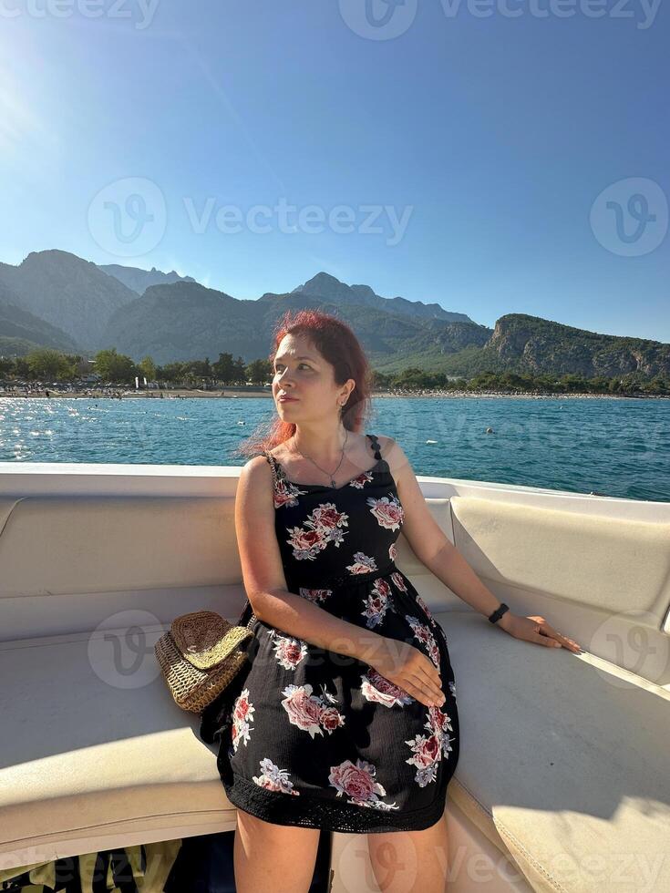 Woman in a black dress sits on a boat under the bright sun, with a scenic view of mountains and the sea in the background. Moment of tranquility and natural beauty concept. Vacation, travel and voyage photo