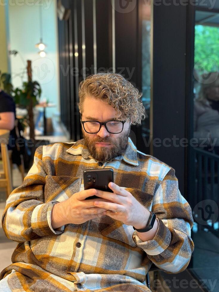 Millennial man with curly hair and glasses is focused on his smartphone while sitting in a cozy cafe. Social networks, communication and technologies concept photo