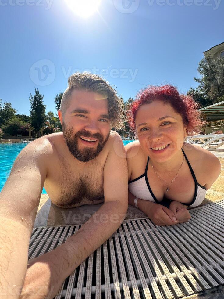 Smiling millennial man and woman takes a selfie while relaxing together at edge of pool on sunny day. Couple enjoy moment in their swimwear sharing happiness and fun. Holidays, vacation and summer photo