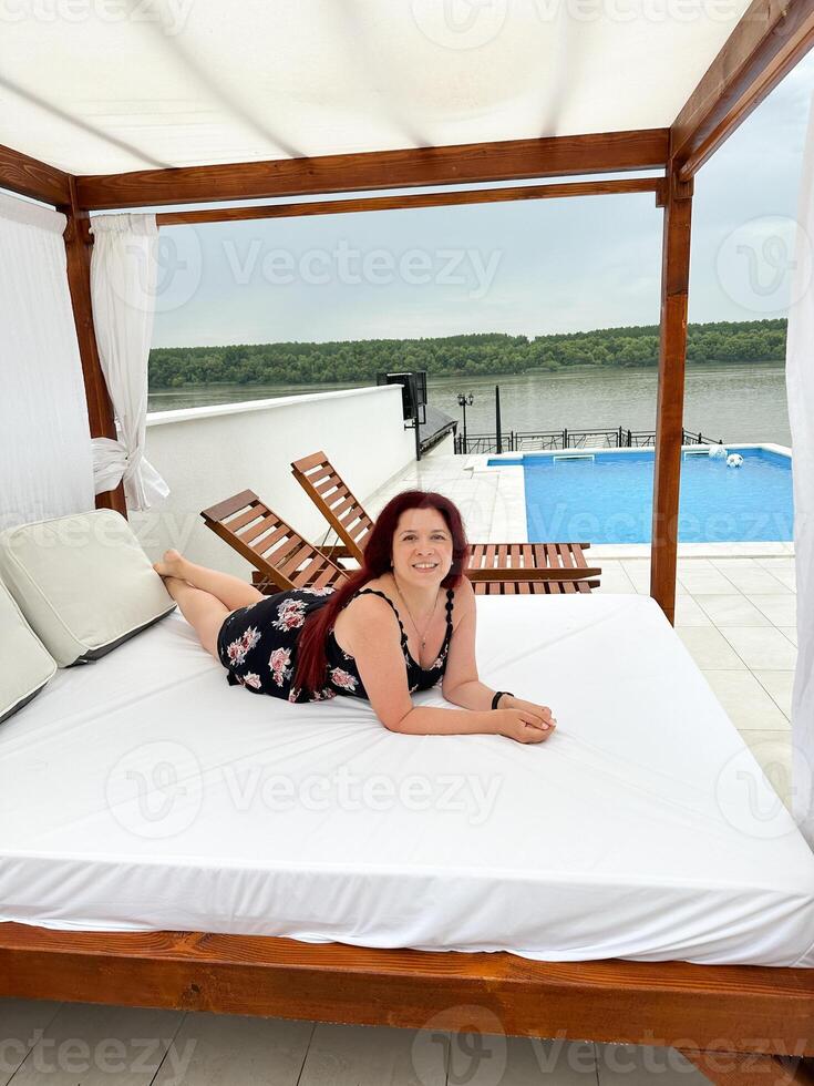 Millennial woman relaxing on a sunbed by a swimming pool, enjoying a serene view. Vacation tranquil setting beach bed and a scenic backdrop of lush greenery. photo