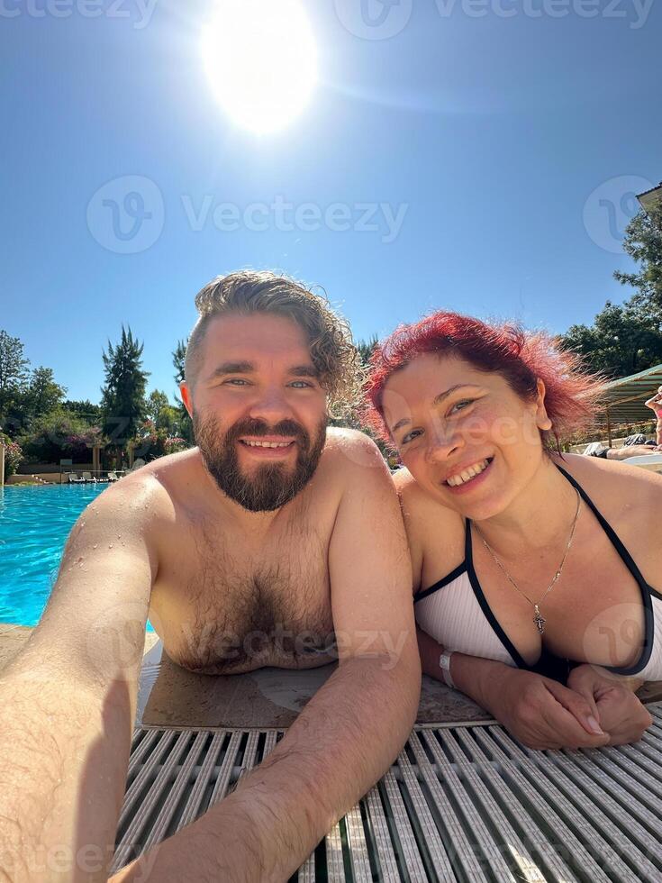 Smiling millennial man and woman takes a selfie while relaxing together at edge of pool on sunny day. Couple enjoy moment in their swimwear sharing happiness and fun. photo