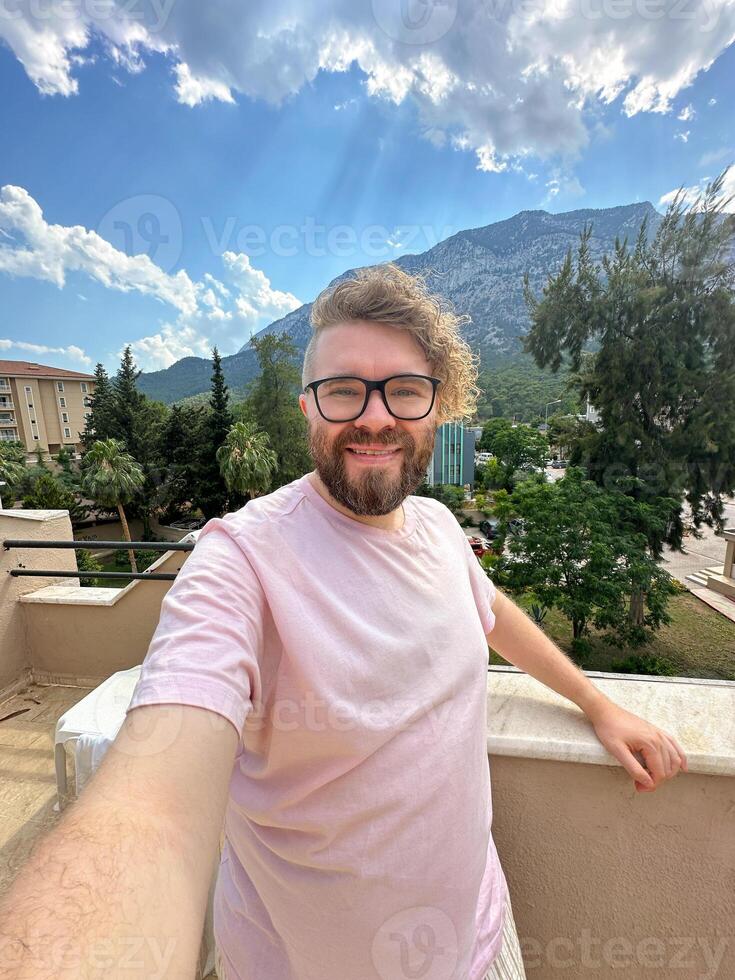 Millennial man with glasses and curly hair is taking a selfie from a scenic terrace. Beautiful mountain landscape under a partly cloudy sky creates a background. photo
