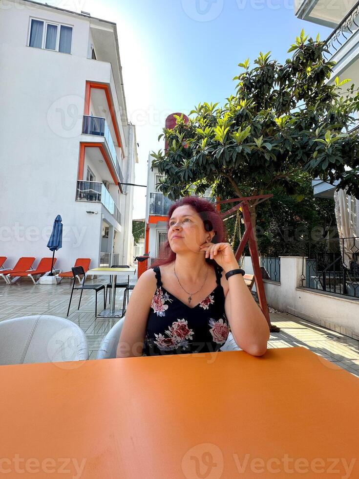 Millennial woman enjoys a sunny day sitting at an outdoor cafe table, surrounded by a modern building and lush greenery. Vacation and summer relax concept photo