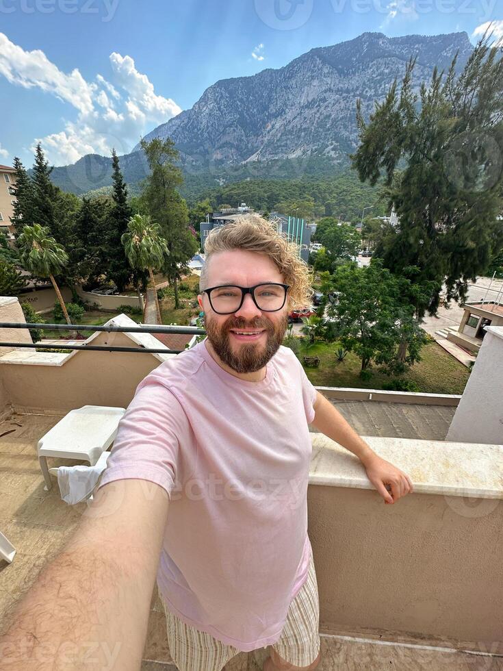 Millennial man with glasses and curly hair is taking a selfie from a scenic terrace. Beautiful mountain landscape under a partly cloudy sky creates a background. photo