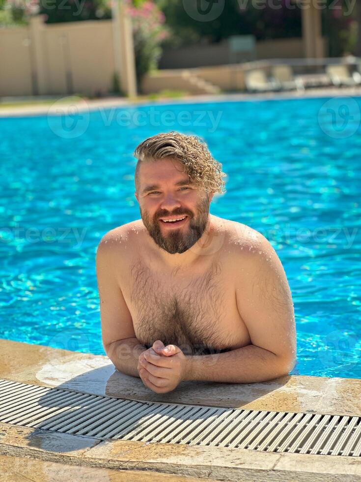 Millennial man enjoying a refreshing day by swimming pool, leaning on the pool's edge and beaming with a broad smile. Holidays and vacation concept photo
