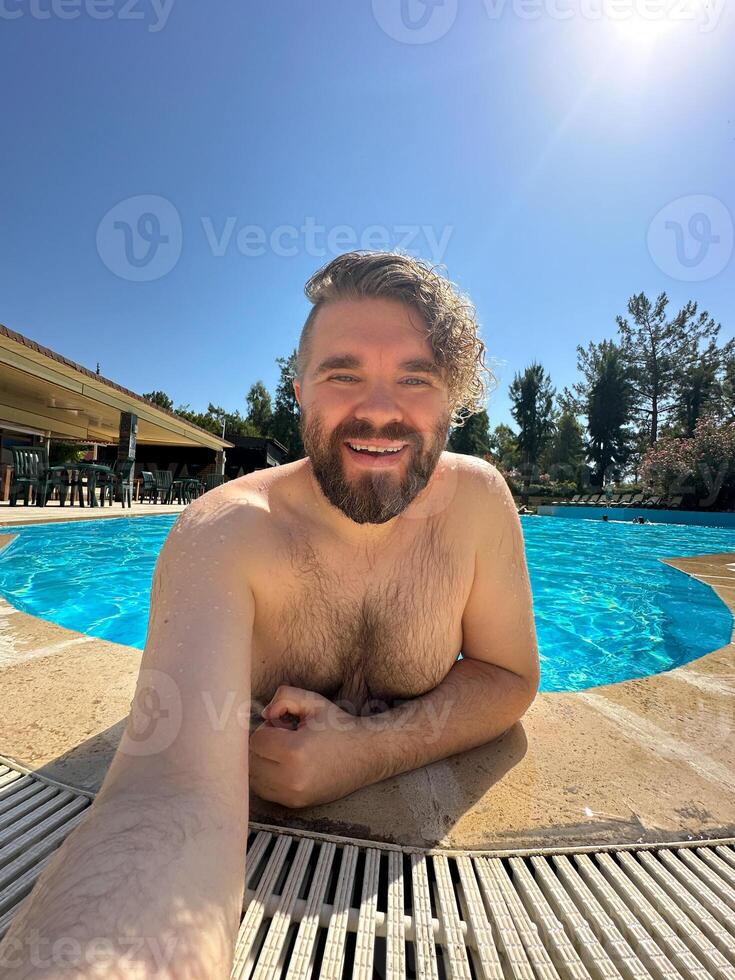 Millennial man taking selfie by swimming pool. Vibrant blue water and relaxed setting create a perfect atmosphere for summer fun. Holidays and vacation concept photo