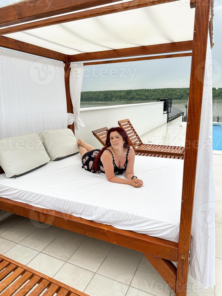 Millennial woman relaxing on a sunbed by a swimming pool, enjoying a serene view. Vacation tranquil setting beach bed and a scenic backdrop of lush greenery. photo