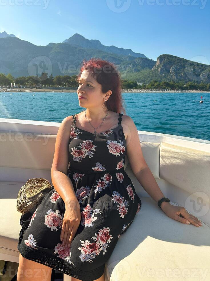 Woman in a black dress sits on a boat under the bright sun, with a scenic view of mountains and the sea in the background. Moment of tranquility and natural beauty concept. Vacation, travel and voyage photo