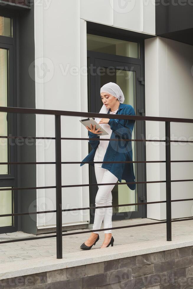 Smiling muslim girl using digital tablet, browsing Internet or social networks outdoor photo