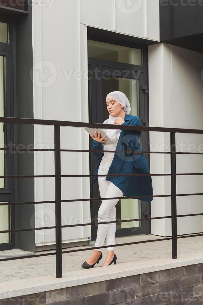 Smiling muslim girl using digital tablet, browsing Internet or social networks outdoor photo
