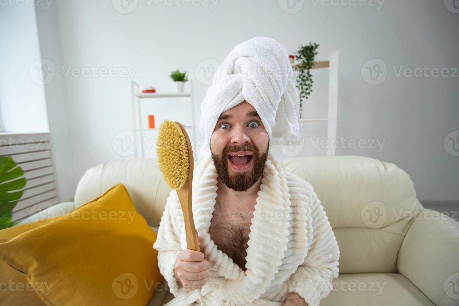 Portrait of funny guy wear turban towel holding in hands massage brush while sits on sofa. Male skin care and spa concept. photo