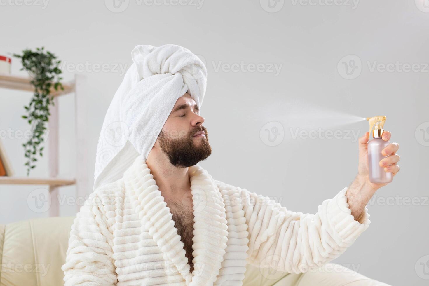 Bearded male with bath towel on his head applying spray water treatment on face. Spa, body and skin care for man concept. photo