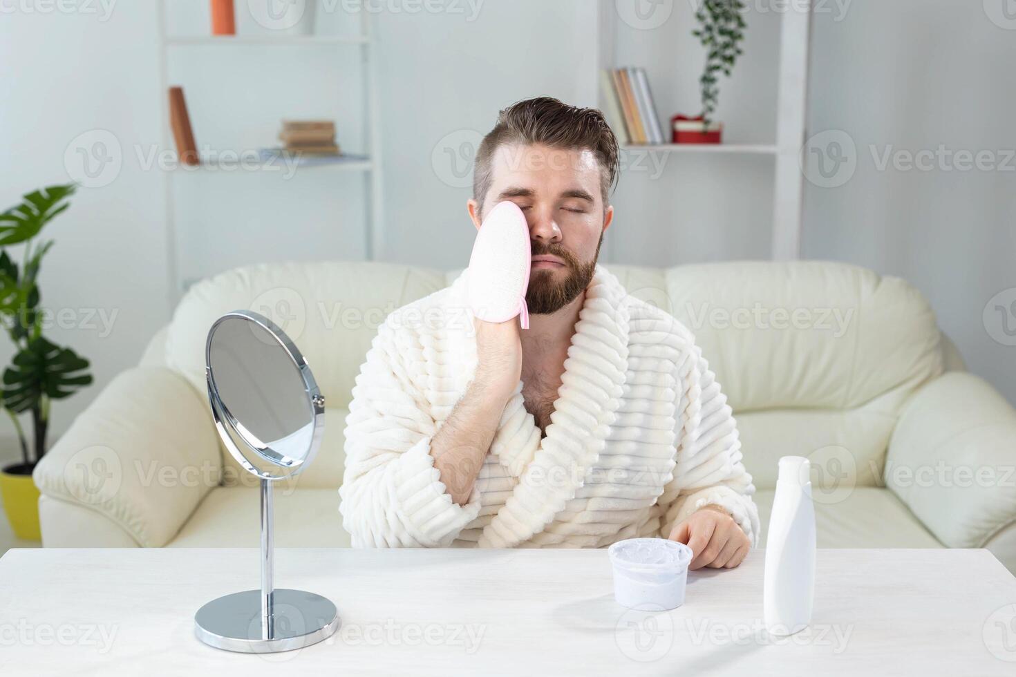 Bearded man look in mirror and clean scrub face with sponge, happy millennial male metrosexual do beauty procedures at home, take care of healthy skin, skincare concept photo
