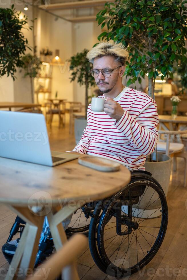Focused guy freelancer with disabilities in wheelchair working online using laptop sits in cozy cafe photo