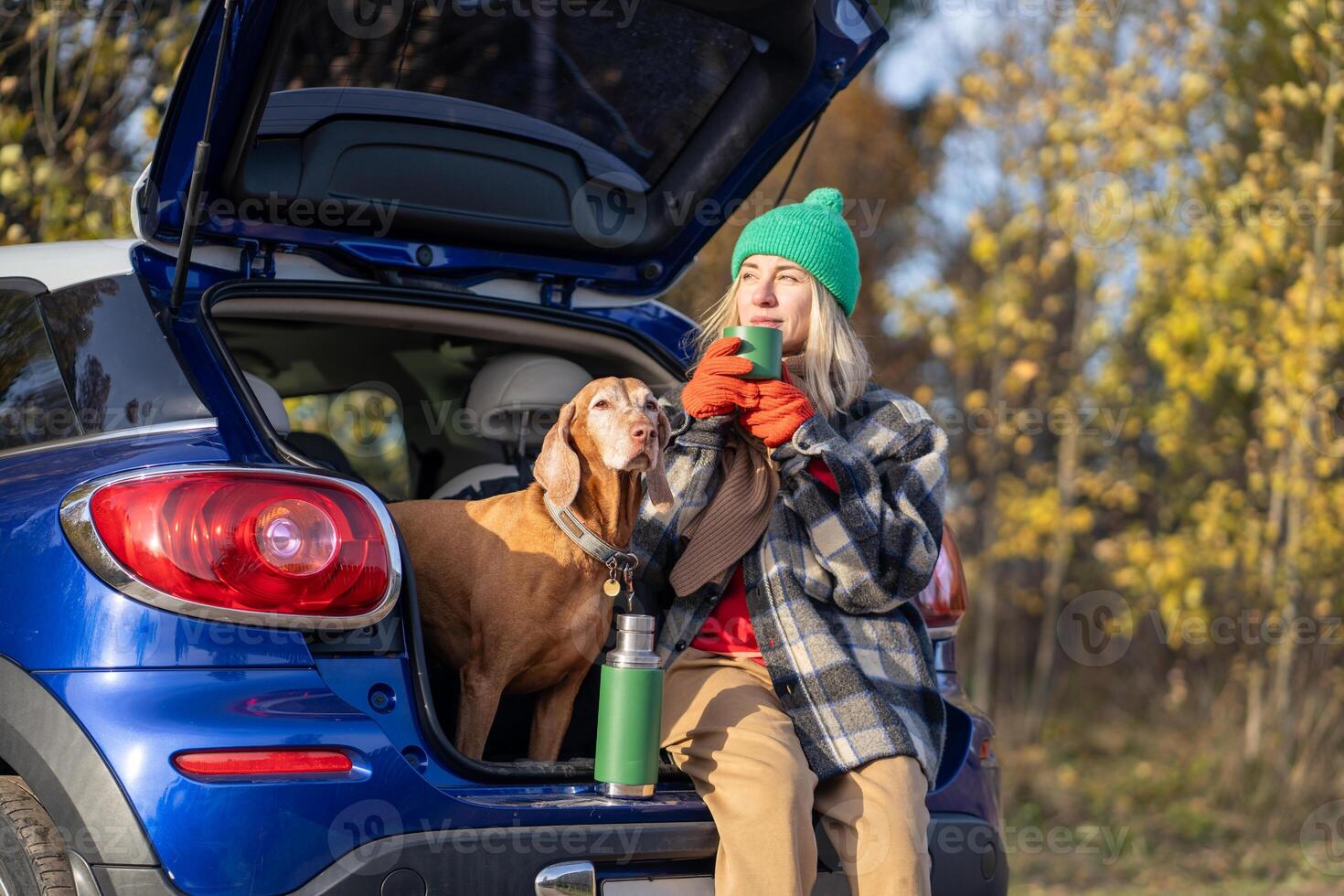 Relaxed woman with dog taking break on road trip, resting with hot tea in car trunk in autumn forest photo