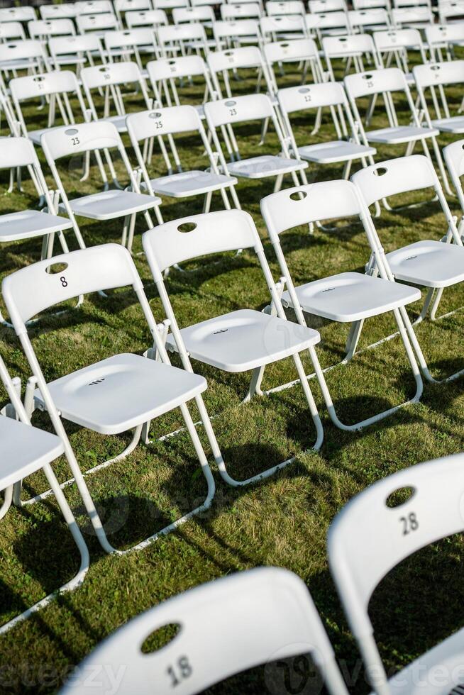 Folding chairs standing in row on green grass, casting shadow on ground. Outdoor party preparation photo
