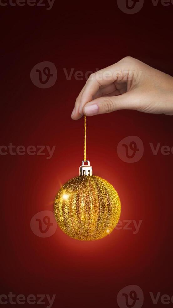 Woman's hand holds a Christmas tree golden ball that rotates. New Year, Christmas minimal concept. Red background copy space vertical banner photo