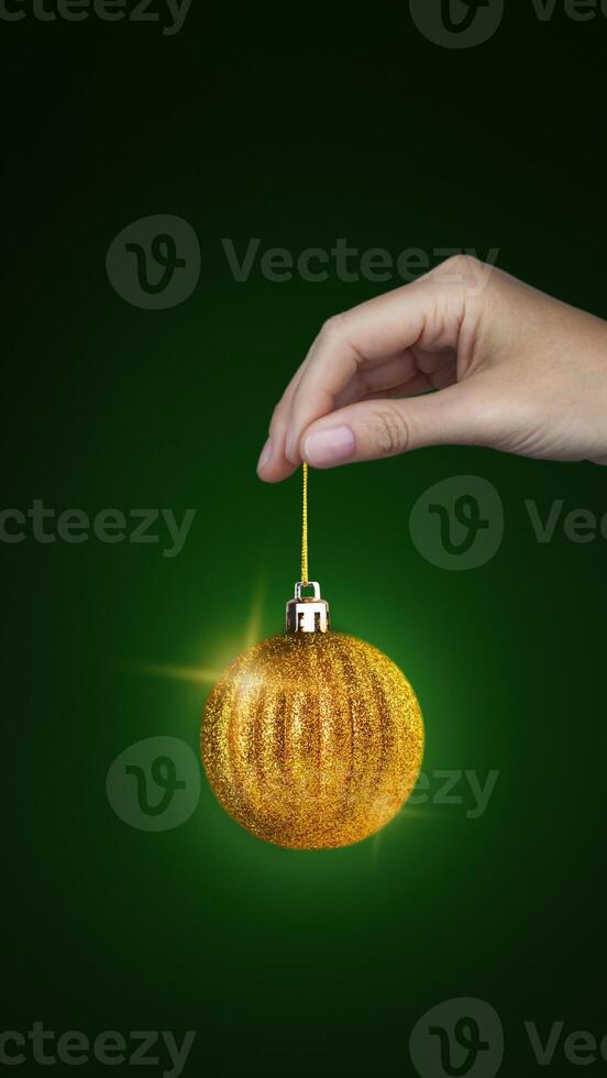 Woman's hand holds a Christmas tree golden ball that rotates. New Year, Christmas minimal concept. Green background copy space vertical banner photo