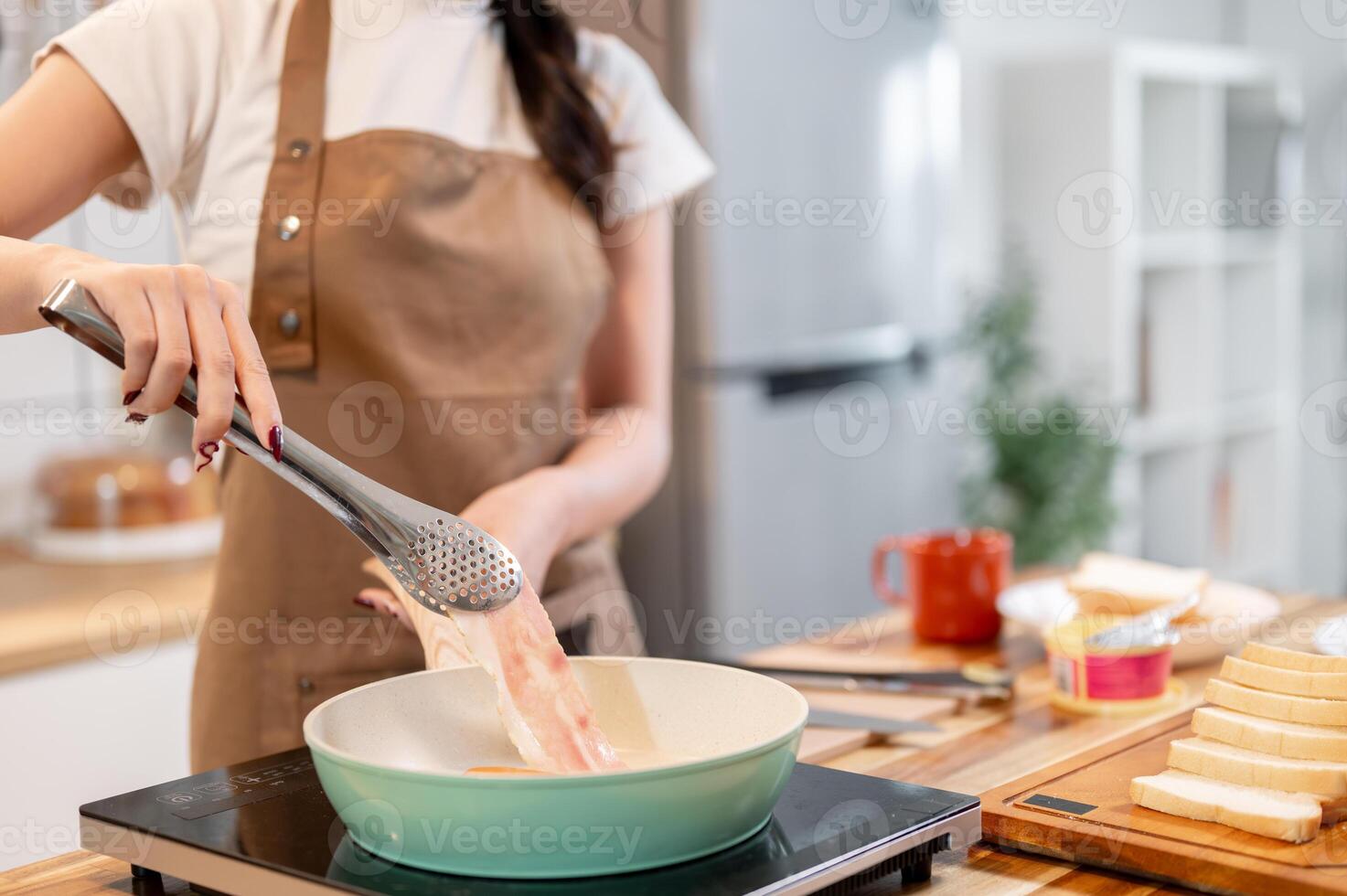 A woman holding tongs, flipping a slice of bacon in a pan while making her breakfast in the kitchen. photo