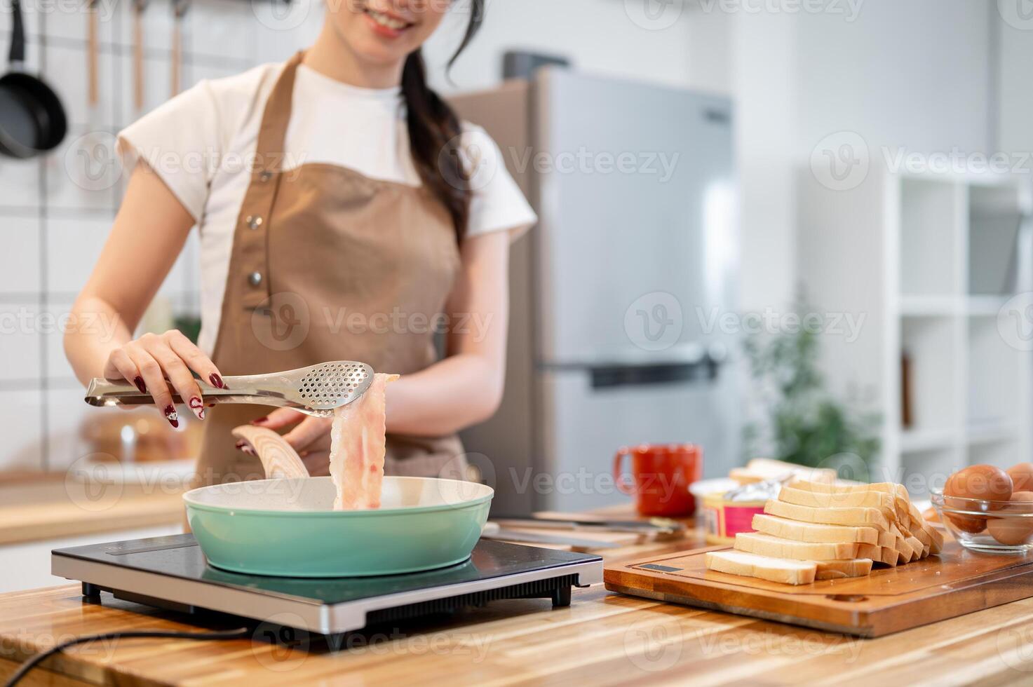 A woman holding tongs, flipping a slice of bacon in a pan while making her breakfast in the kitchen. photo