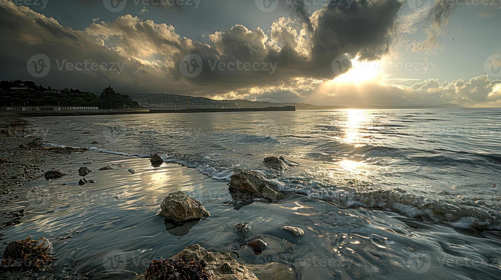 A dramatic sunset over a calm sea with large, dark clouds, the sun shining through and reflecting off the water. There are a few rocks in the foreground and a small wave is breaking on the beach. photo