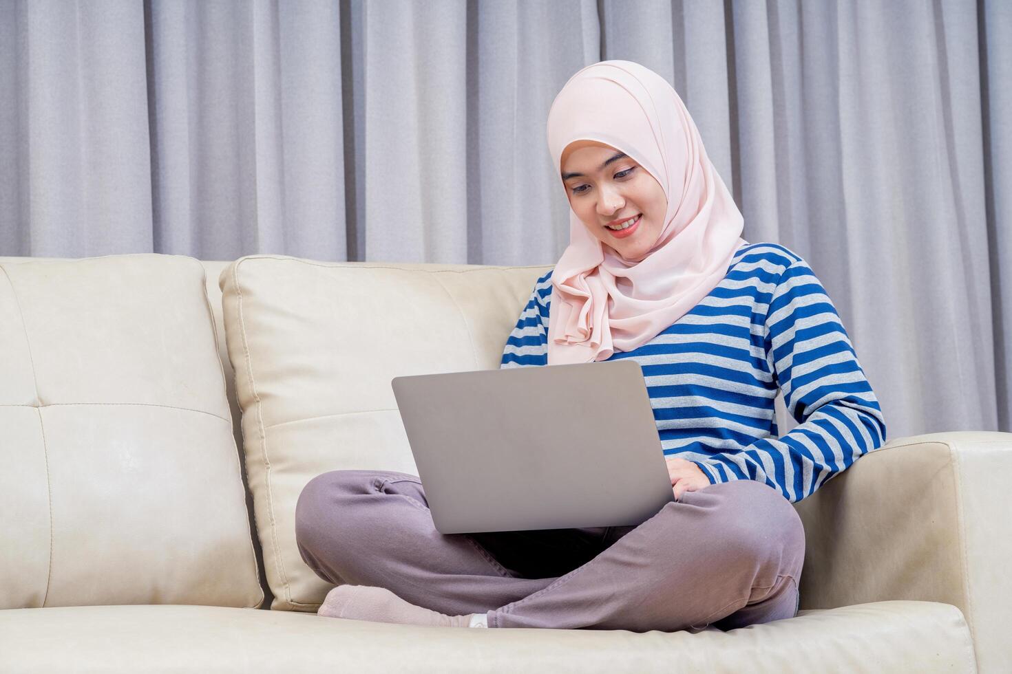 Asian muslim woman wearing hijab sitting using the laptop to do work, working from home at her house on the sofa and sit and watch television. photo