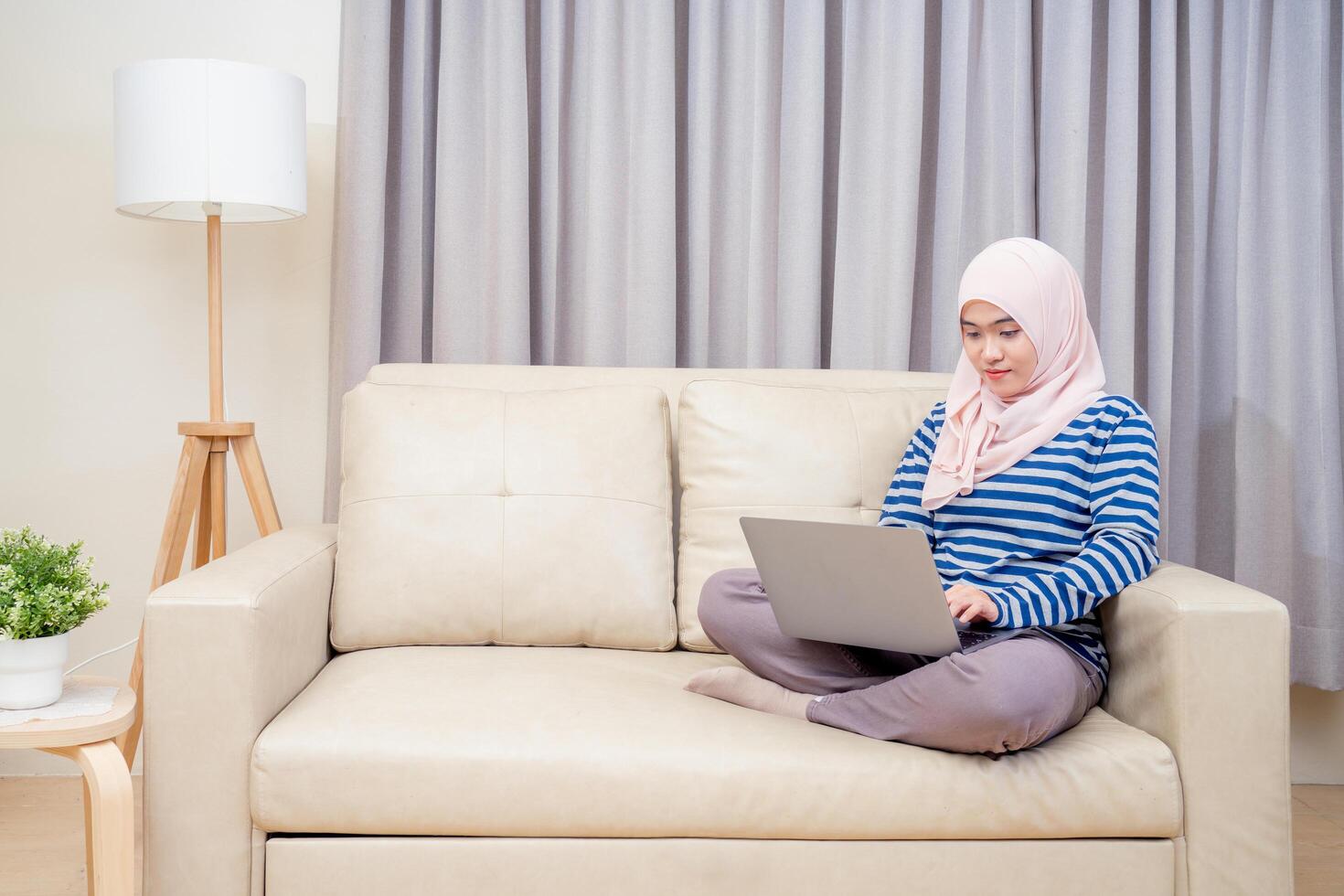 Asian muslim woman wearing hijab sitting using the laptop to do work, working from home at her house on the sofa and sit and watch television. photo